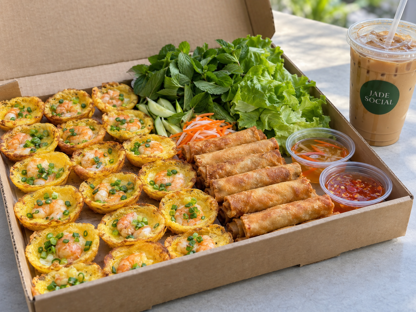 Open shallow kraft catering box with rows of golden shrimp bánh khọt and Vietnamese egg rolls, fresh herbs, sauces and iced coffee